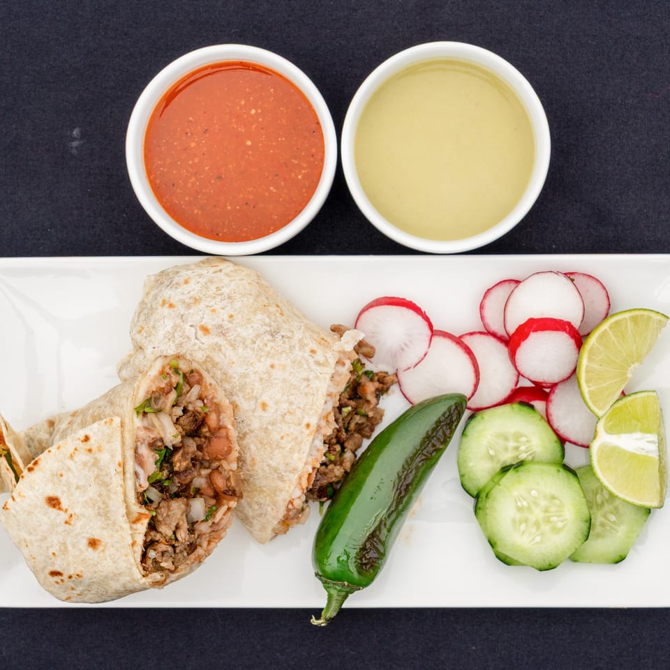 Two Blady's Tacos burritos with sides of sliced radishes, cucumbers, lime, a jalapeño, and two dipping sauces.