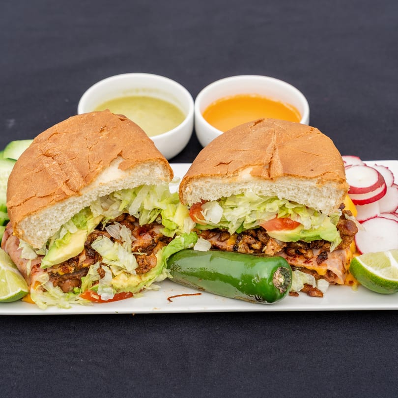 Two Blady's Tacos tortas with meat, lettuce, avocado, and tomato, with sauces, radishes, and lime.