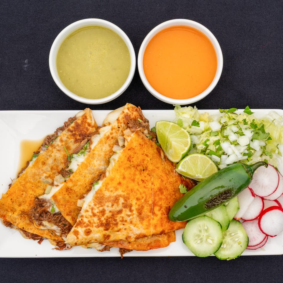Blady's Tacos birria quesadillas with two dipping sauces, limes, jalapeño, and fresh vegetables.